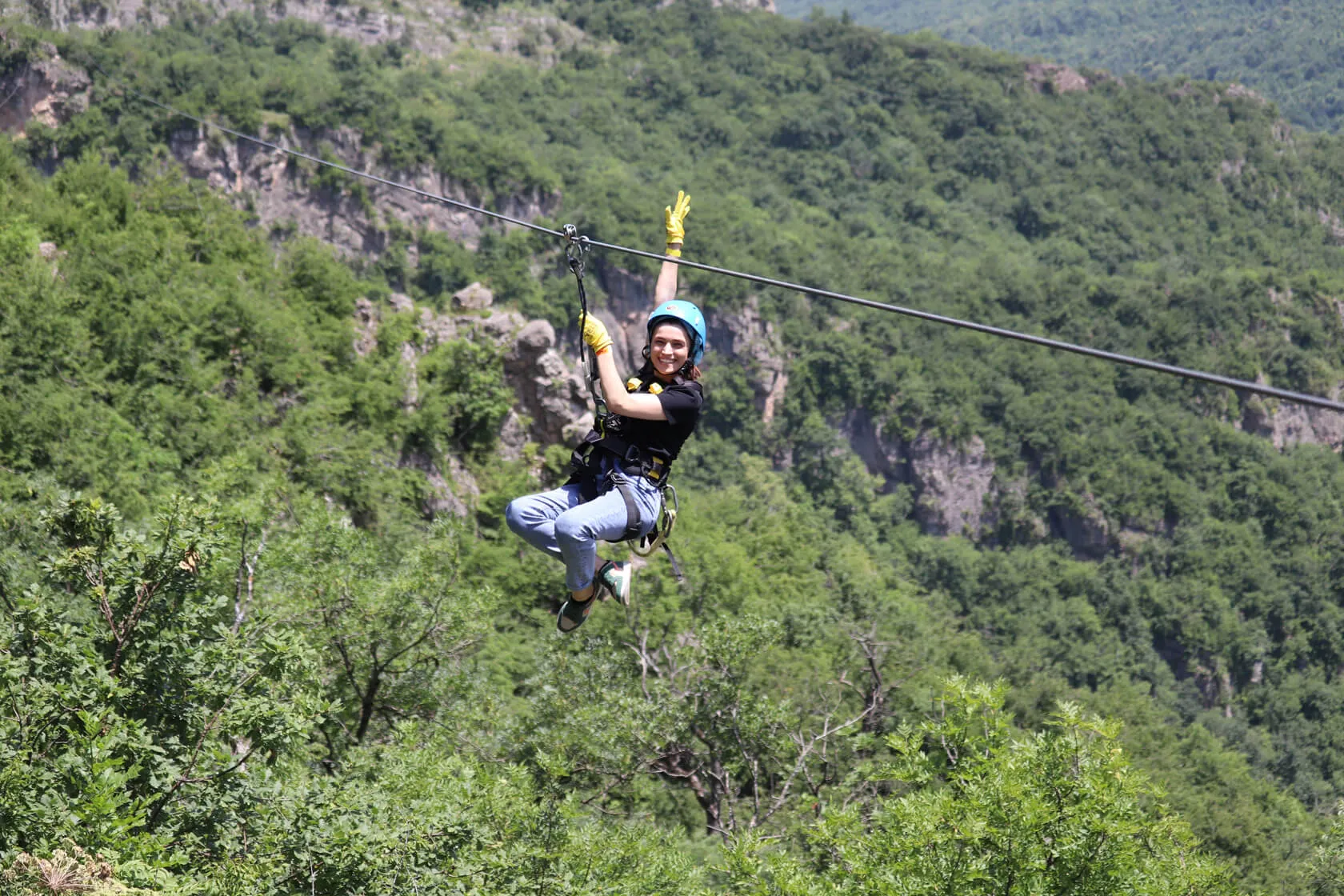 Rider in a blue helmet waving from the zipline above the forested canyon